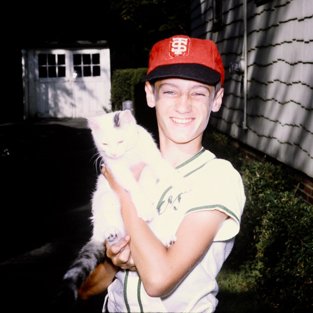 Dan around age 12, wearing a red cap with a black peak and a white baseball jersey with green piping while holding a white cat.