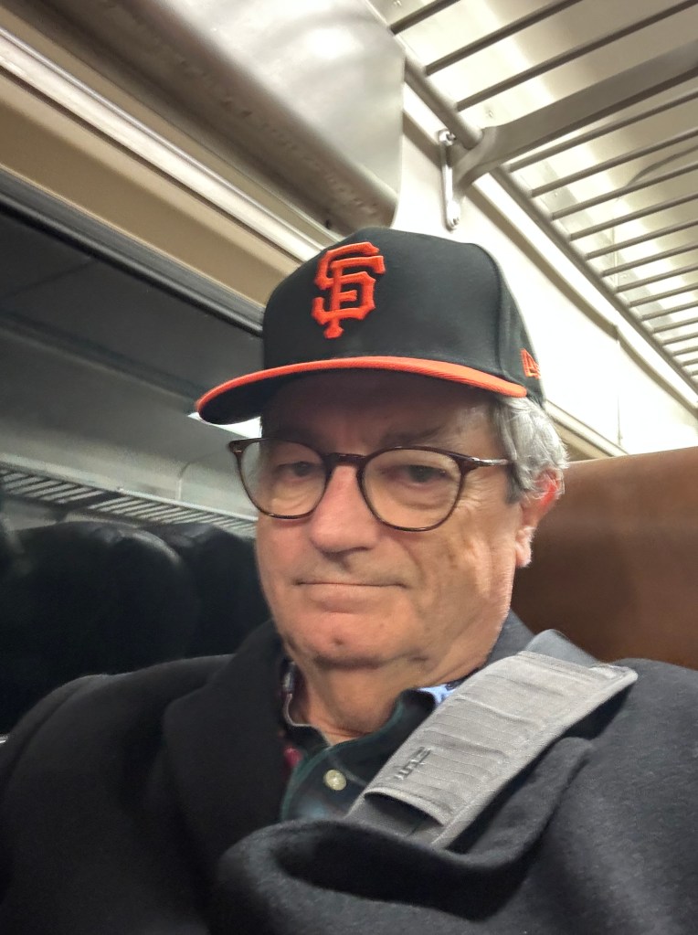 The author sitting in a New Jersey Transit train car. He is wearing a San Francisco Giants cap, and a padded shoulder strap is on his shoulder, over a black wool coat.