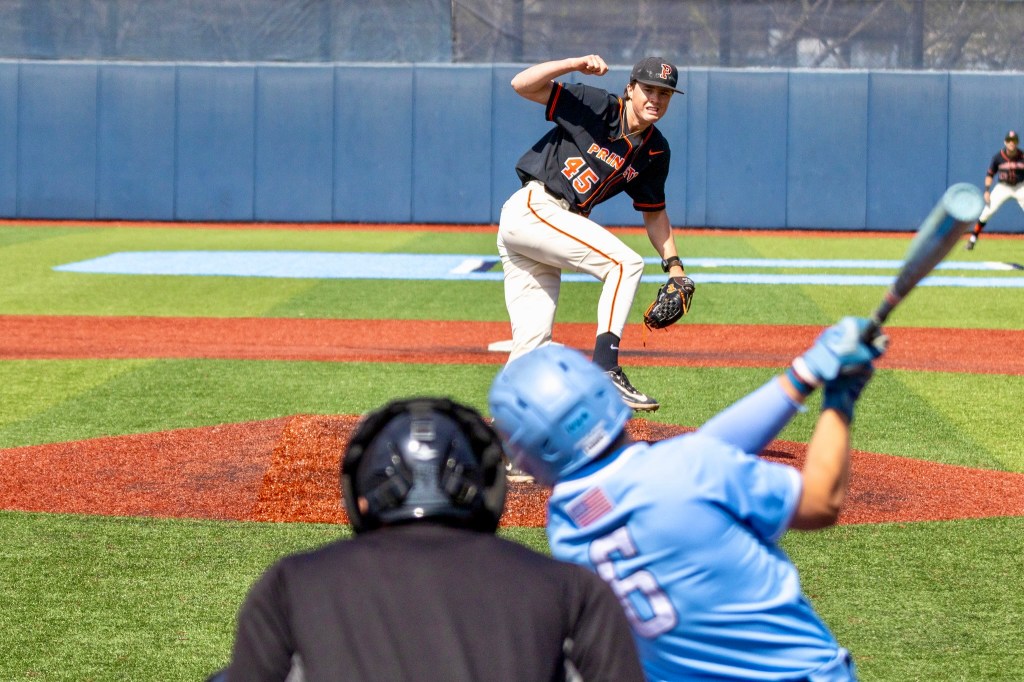 Princeton pitcher strikes a pose with right arm curled high after completing delivery of a pitch to the Columbia batter, who swings past the ball. The umpire looks on from behind.

