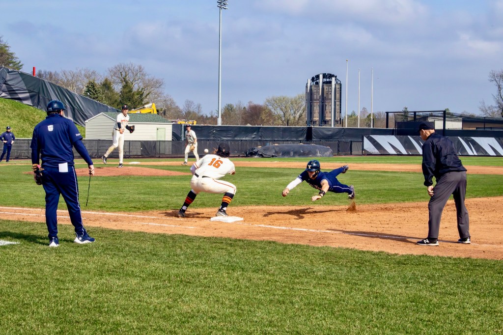 Yale baserunner dives to first base as a pickoff throw from the pitcher is a split seond away from entering the first baseman's mitt. The umpire and the Yale first base coach look on, framing the action at the base.