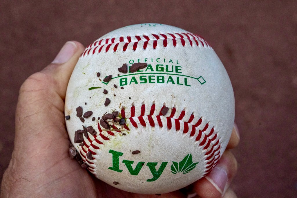 Hand holding a dirt-flecked baseball bearing the Ivy League logo in green.
