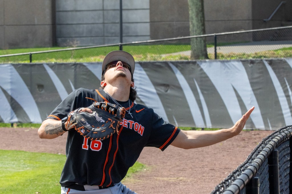 Princeton first baseman stick his left arm out to find the baseline fence as he raises his mitt to try to catch a foul ball going out of play.