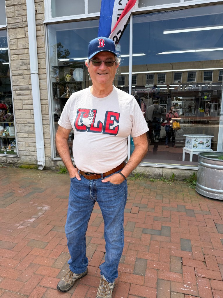 Man wearing a Boston Red Sox baseball cap, blue jeans and a white Cleveland Guardians T-shirt with CLE in bold letters across the chest. He is standing on a brick sidewalk with an antique store behind him.