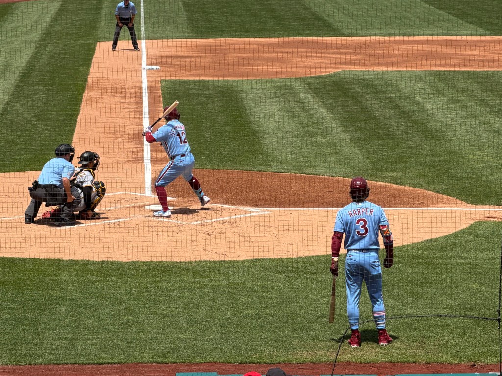 Kyle Schwarber lifts his front foot as he anticipates the arrival of the pitch. Bryce Harper, holding his bat along his left leg, looks on. The Phillies were in power blue uniforms with maroon caps.