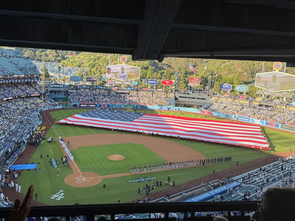 A huge American flag stretches across the outfield. The Dodgers players line the third base line, and the Brewers are lined up on the first base line.
